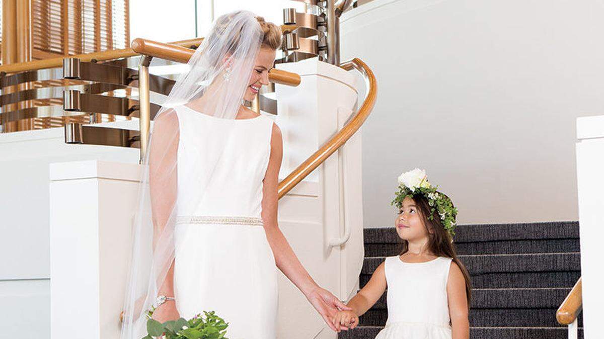 GRAND ENTRANCE On bride: Modern Trousseau “Minnie” dress with a bateau neckline, V back and chapel train, $3,400, and beaded Finley belt, $358, Sara Gabriel “Matty”earrings, $197, and “Daphne” bracelet, $257, all from Altar Bridal. On flower girl: Rosebud dress with scoop neckline and pearl-trimmed waist, $118, from Bella Bridesmaids. Bridal bouquet and flower crown by Beco Flowers