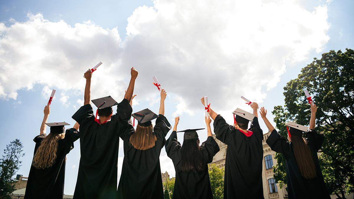 Rear portrait of group students raise fists hold diploma mortarboard gown graduate college building outdoors. high school graduation