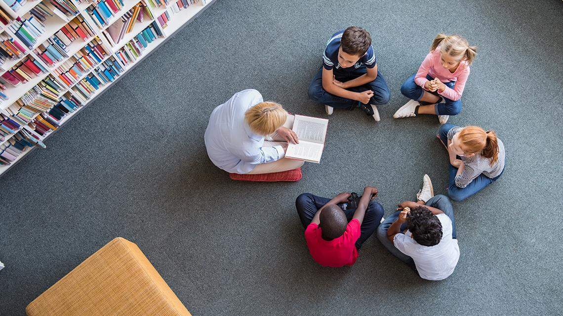 Teacher reading fairy tales to children sitting in a circle at library. Top view of librarian sitting with five multiethnic children on floor. Teacher reading book to girls and young boys at school.