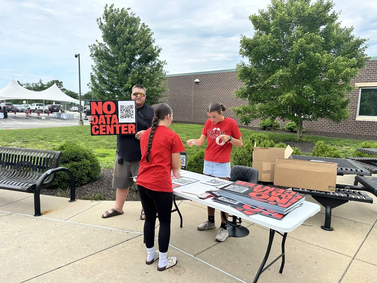 Lawrence resident Sterling Ozark holds up a “No Data Center” sign alongside two De Soto High School students outside of Beale’s Community Open House on June 18, 2026. Ozark was handing out yard signs that include a QR code to De Soto’s data center opposition Facebook group.