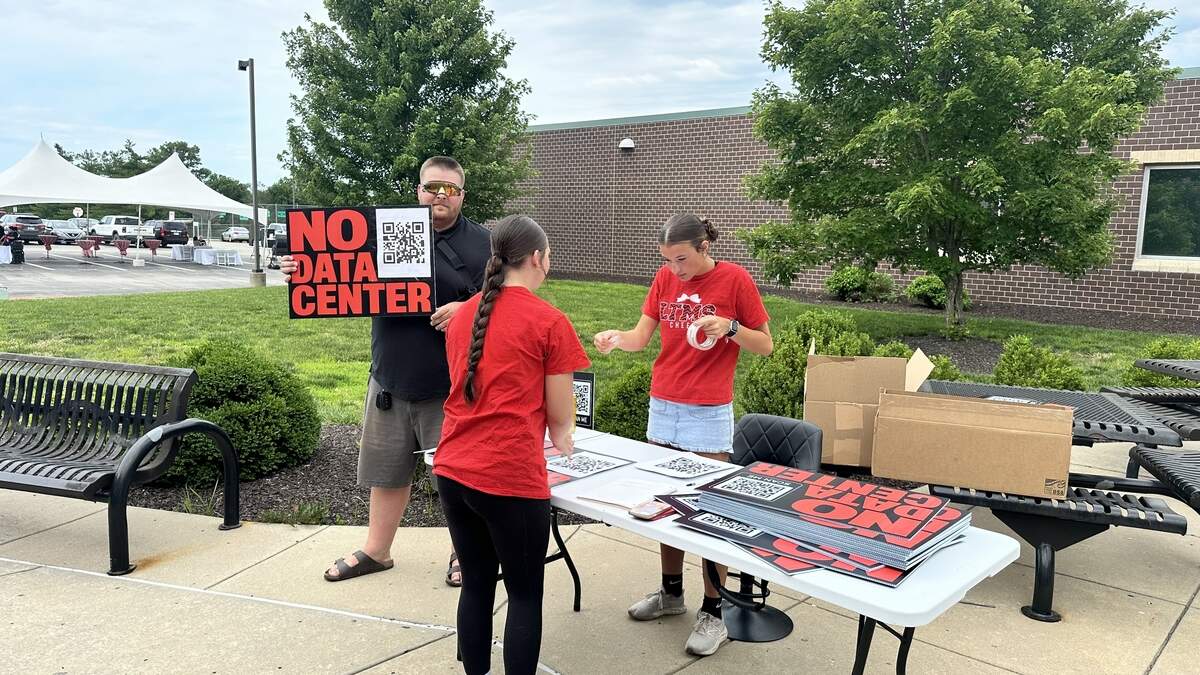 Lawrence resident Sterling Ozark holds up a “No Data Center” sign alongside two De Soto High School students outside of Beale’s Community Open House on June 18, 2026. Ozark was handing out yard signs that include a QR code to De Soto’s data center opposition Facebook group.