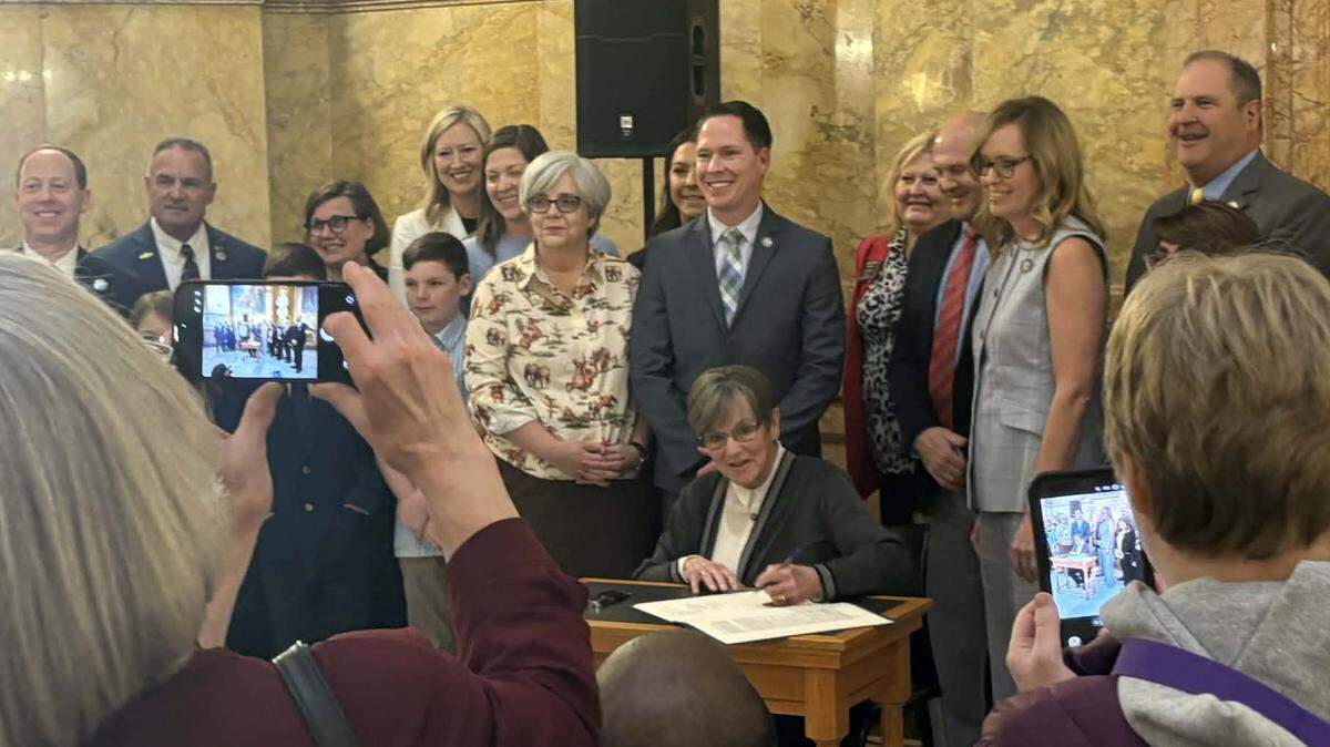 Kansas Gov. Laura Kelly signs a bipartisan school cellphone ban bill into law on March 19, 2026 at the Statehouse as onlookers snap cellphone photos.