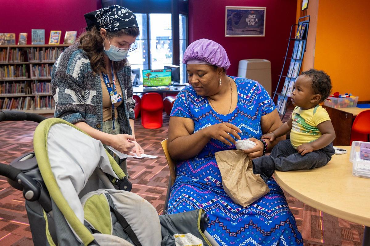 Senior Library Assistant Clyde Miedema, left, chats with Zeylona Burnett, who holds her 11-month-old son, Kyrie, and a free sack lunch, as they discuss a list of free meal locations from the Free Hot Soup organization on at the Lucile H. Bluford Library.