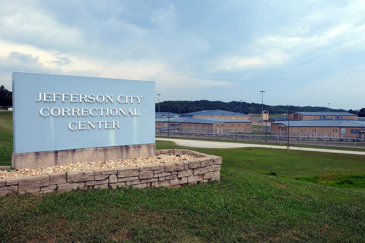 An image shows a light blue sign that reads “Jefferson City Correctional Center.” Gray and tan prison buildings surrounded by wired fences are in the background.
