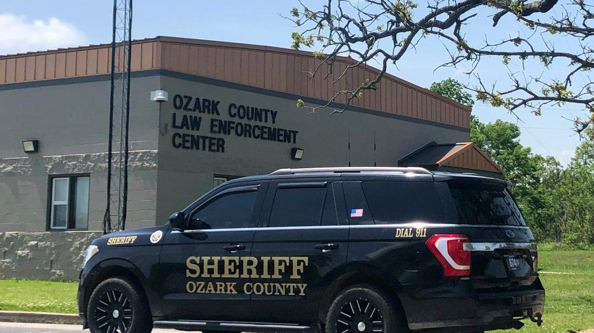 A Black SUV with the words “Sheriff Ozark County” on the driver’s door is parked in front of a gray brick building with a sign that reads “Ozark County Law Enforcement Center.” 