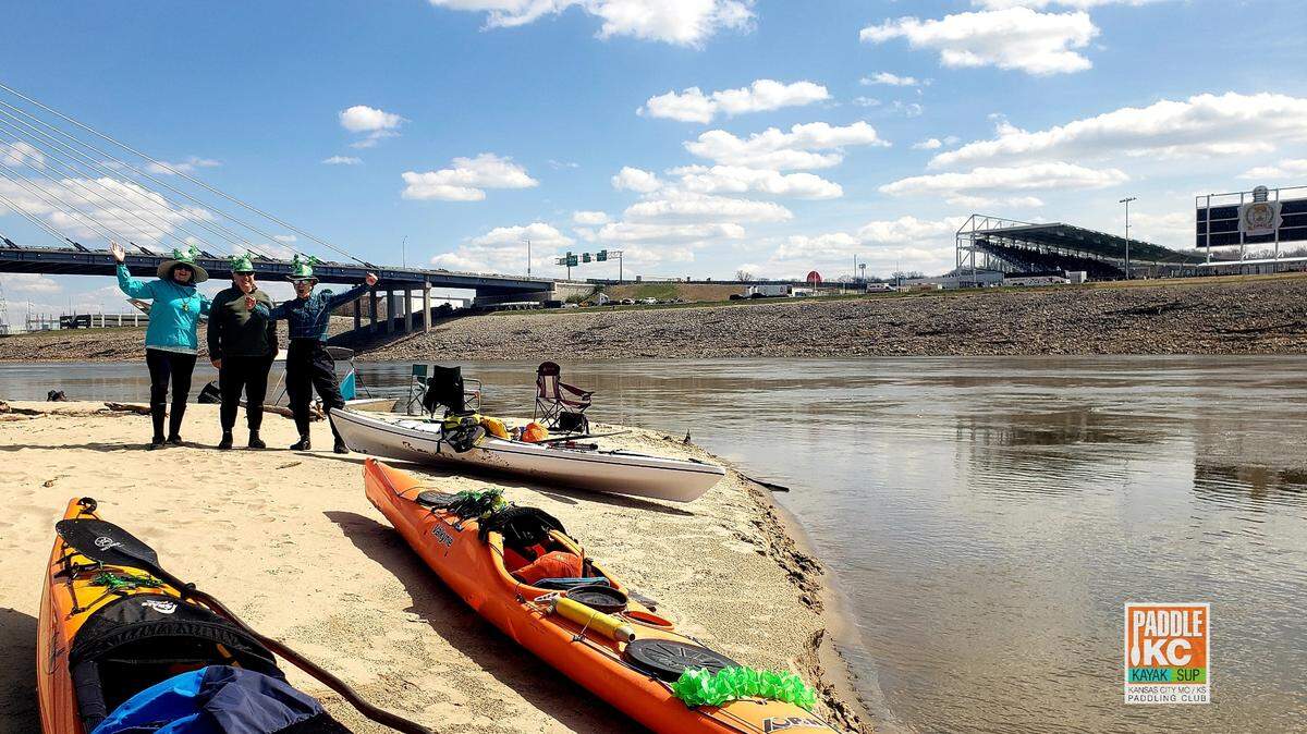 Paddlers and their kayaks on a sandbar of the Missouri River during the KC Current’s season opener at CPKC Stadium (background) in Kansas City.