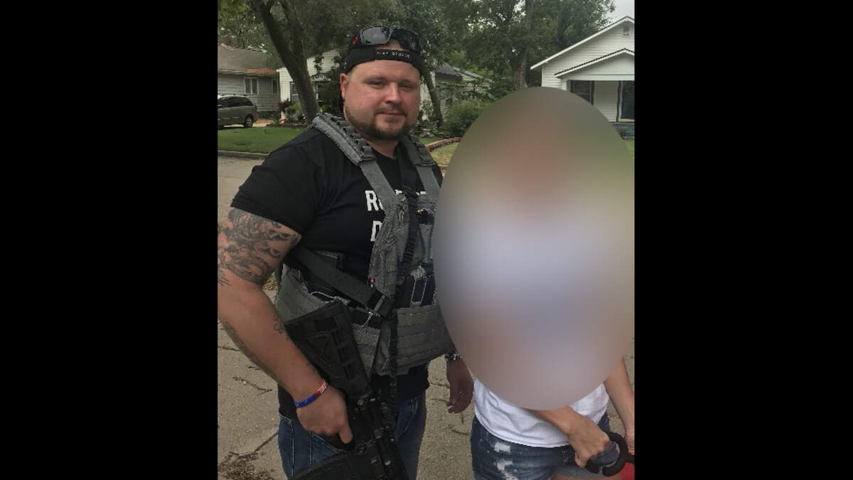 Michael Eckerman, holding an assault rifle and wearing a tactical vest, stands next to his wife.