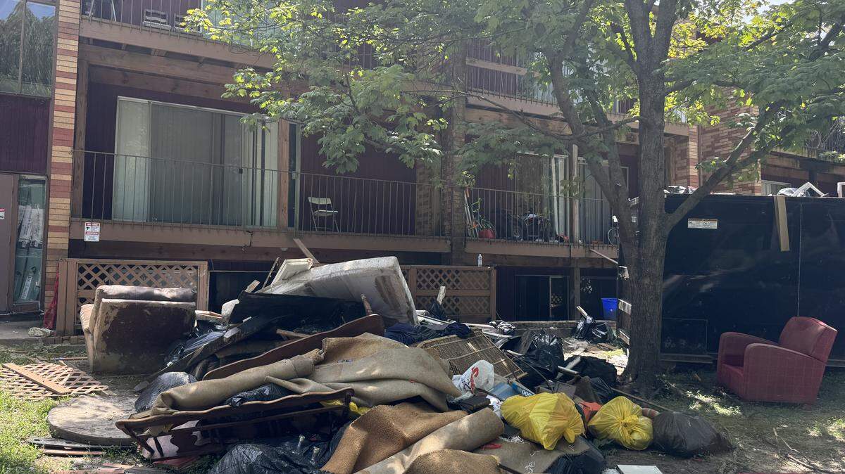 Outside the apartments is a pile of ruined belongings and furniture and a dumpster.