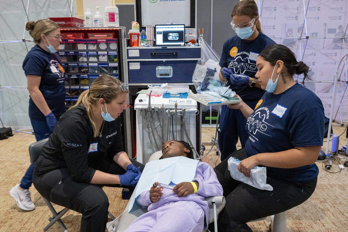 TeamSmile volunteers looked on as Caitlin Chidsey, left, a pediatric dentist from Olathe, left, talks to Khloe Harris, 7, of Kansas City, who was getting dental care from the Kansas City Chiefs and TeamSmile, who sponsored a dental tailgate to provide free dental care to 300 school children from Kansas City, Missouri and Kansas City, Kansas, on Tuesday, Sept. 9, 2025 at GEHA Field at Arrowhead Stadium.