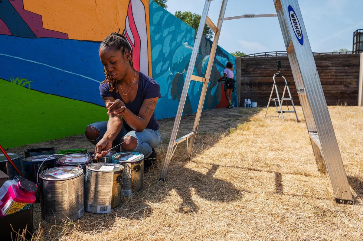 Jasmine Ali, left, opens up paint cans to continue painting her mural while Emily Alvarez finishes her art piece on Friday, Sept. 15, 2023, at the Riverfront Heritage Trail in Kansas City. Ali has worked as a professional artist for seven years and is part of the first group of artists painting murals on the concrete wall.