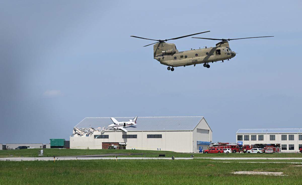 A Chinook helicopter makes a landing while a Beechcraft airplane rests on the roof of a building at New Century AirCenter after it made a crash landing that left it on the roof on Monday, June 16, 2025.