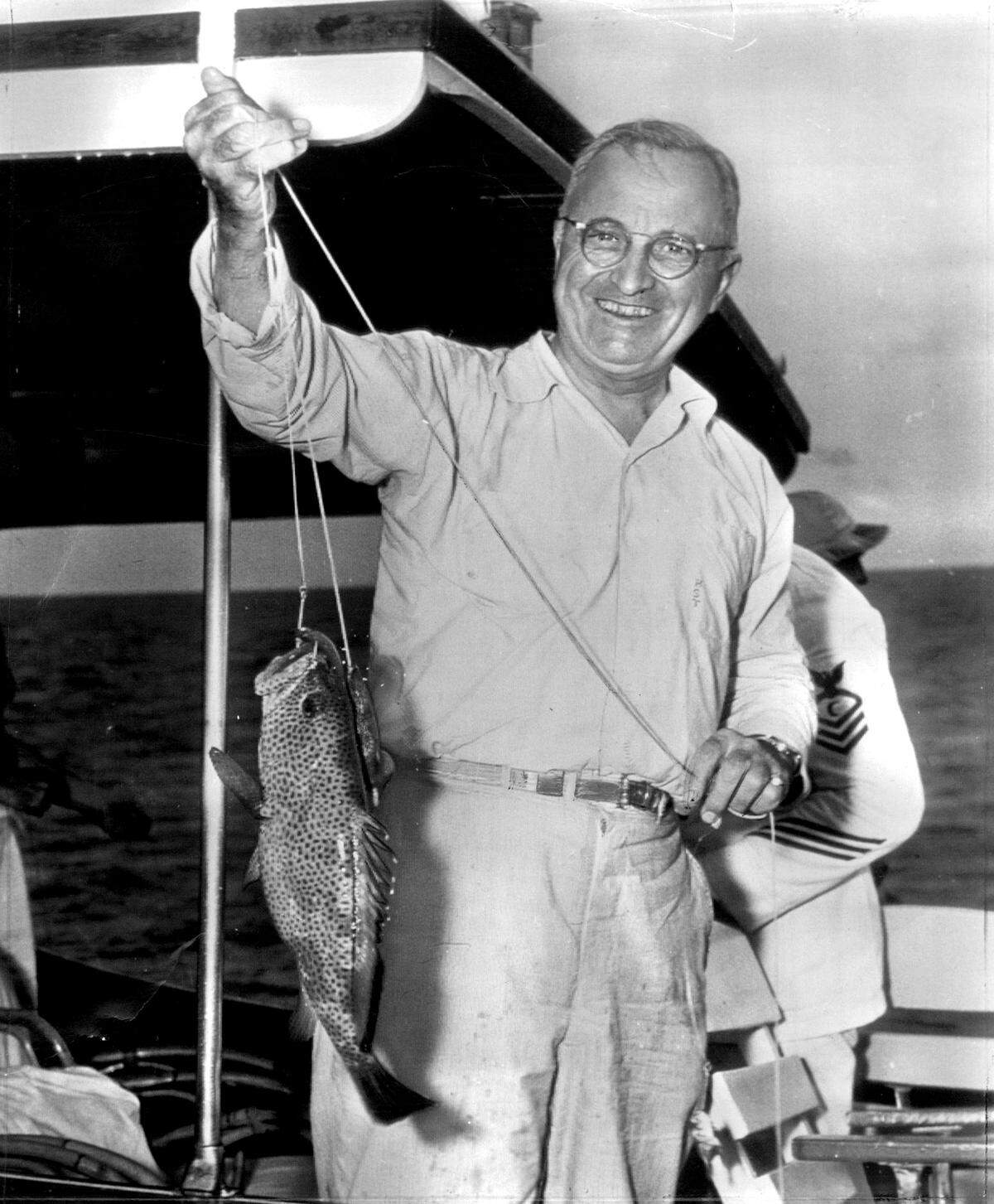 President Harry S. Truman holds up a fish he caught during a 1946 vacation cruise to Bermuda. He was in a small boat when the photo was taken, with a Navy cook partially visible behind him.