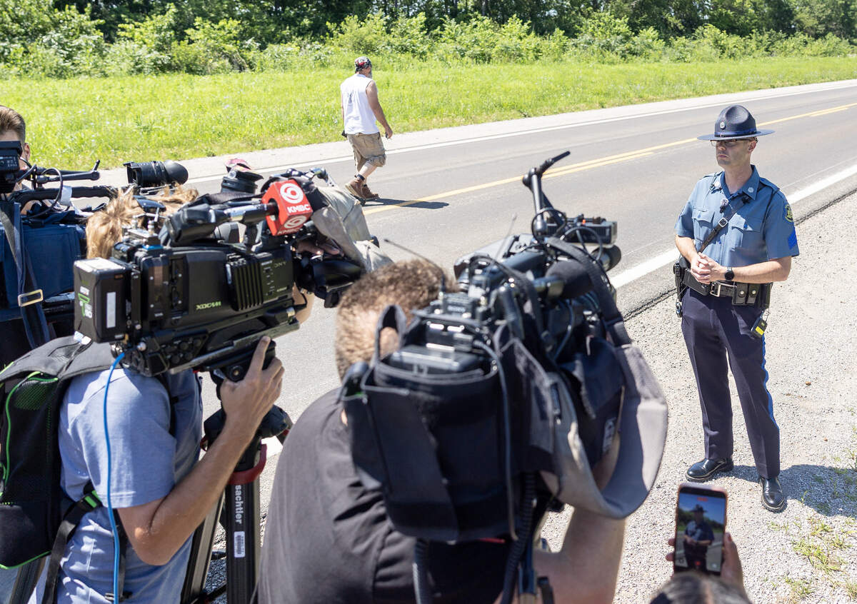 Sgt. Justin Ewing, a spokesperson for the Missouri State Highway Patrol, speaks to the media about a plane crash in Butler, Missouri, on Sunday, June 14, 2026, as a man who identified himself as a family member of a person onboard the plane walks toward a family notification area near the plane crash site. The man declined to give his name.