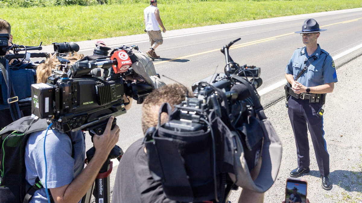 Sgt. Justin Ewing, a spokesperson for the Missouri State Highway Patrol, speaks to the media about a plane crash in Butler, Missouri, on Sunday, June 14, as a man who identified himself as a family member of a person onboard the plane walks toward a family notification area near the plane crash site. The man declined to give his name.