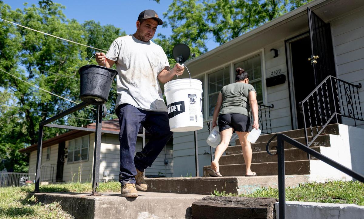 Alfonso López walks with empty buckets to retrieve water from his car as his wife Jessica Martinez carries jugs of water inside on Friday, May 26, 2023, in Kansas City. Due to an outstanding balance left by a previous tenant, KC Water shut off water to the couple’s home in April. Since then, their family has had to get creative for their water needs including transporting old pop bottles and buckets full of water by car to their home and using bottled water to brush their teeth.