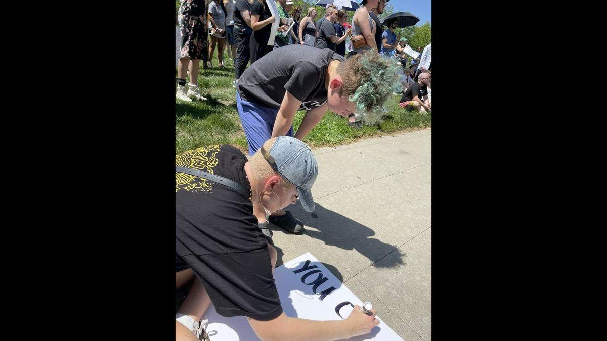 Cassie Taylor, 36, and their child Woods Haren, 8, designed a poster in the crowd of people gathered Saturday in Ilus Park to protest restrictions on gender-affirming care. Taylor wrote “You cannot make us disappear” in bold letters.