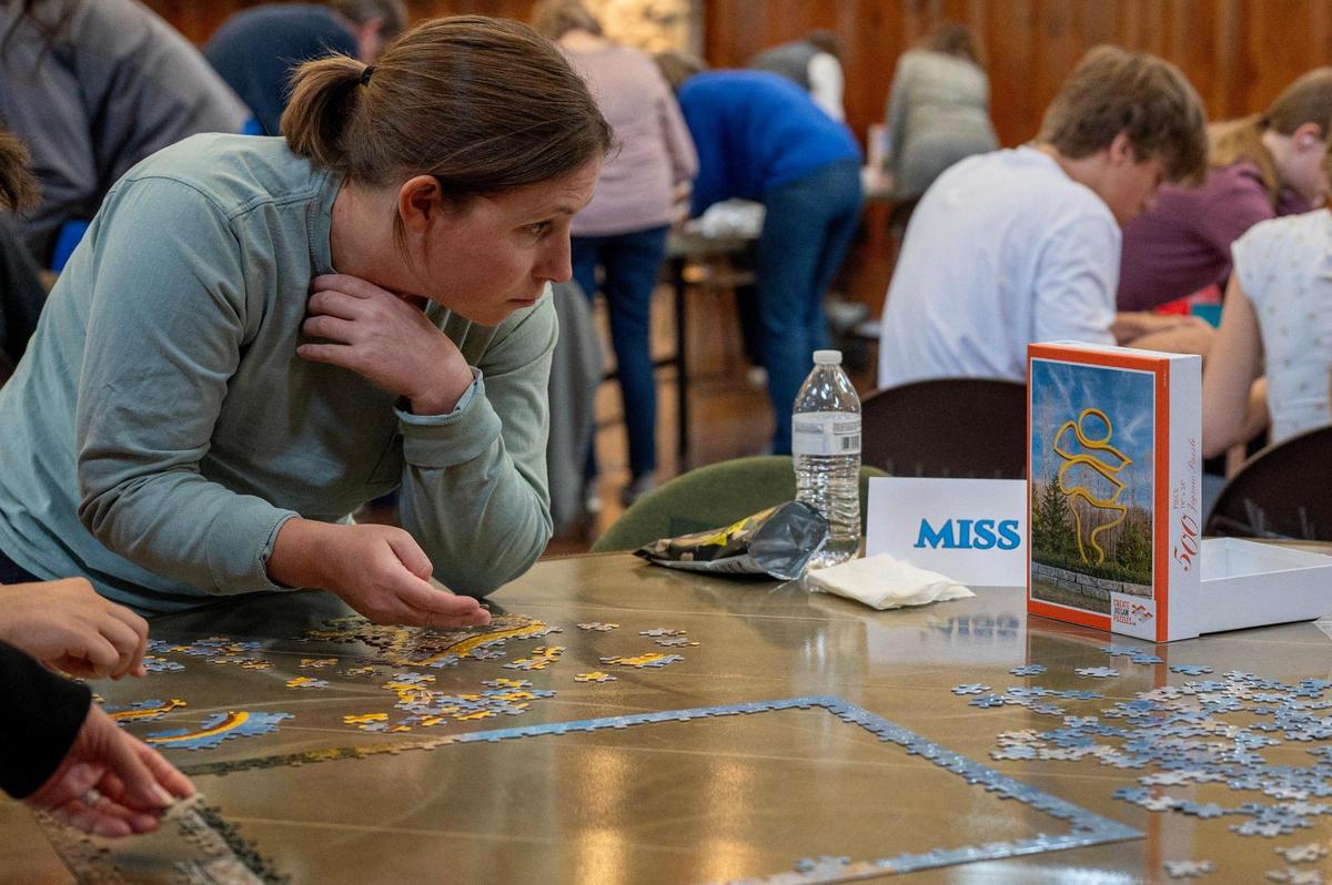 A team member from Miss Fits compares puzzle pieces to the reference image on the box while working with their team to complete a 500-piece puzzle during the Puzzle Palooza competition.