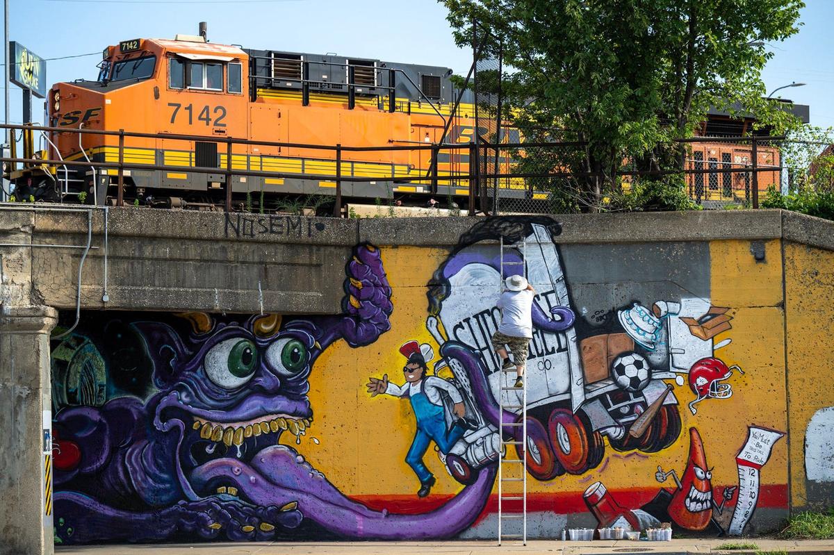 A freight train passes by as artist Mike Elder climbs a ladder to paint a section of his mural on the Independence Avenue truck-eating bridge.. Elder’s mural features a purple troll attacking a truck, spilling its Kansas City-themed cargo.
