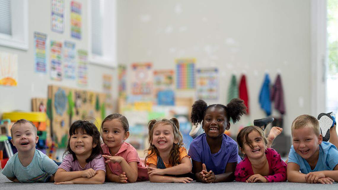 A small group of Kindergarten students lay on the floor of their classroom, side-by-side, as they pose for a portrait.  They are each dressed casually and are smiling.