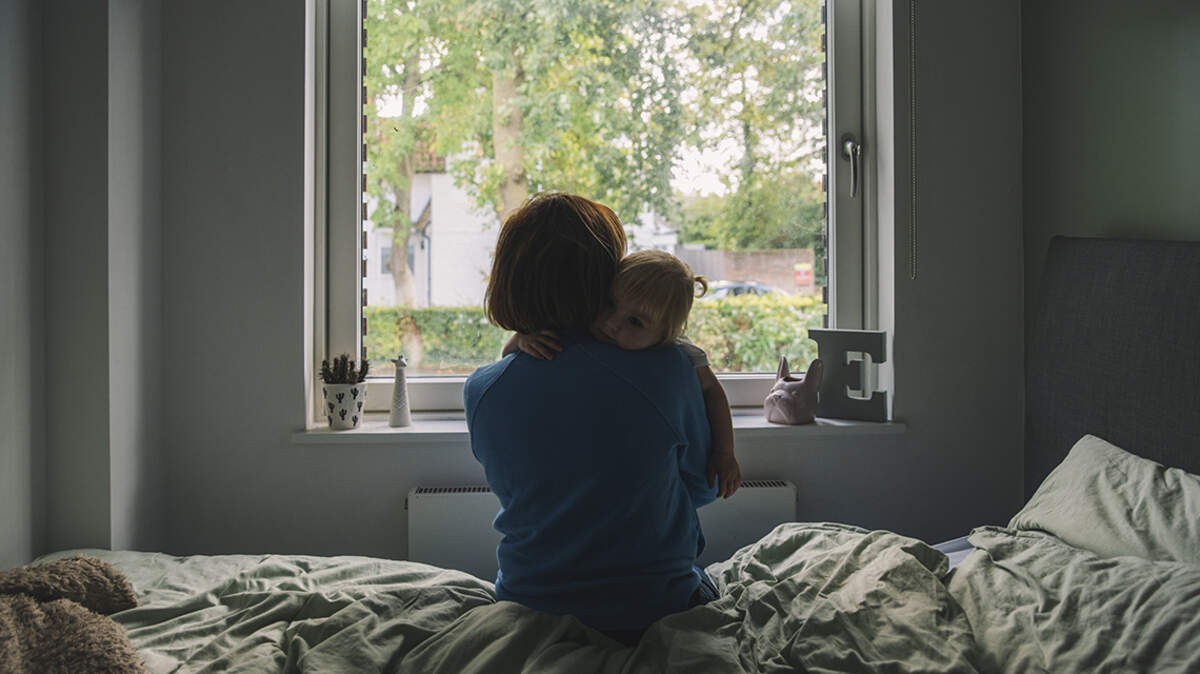 A woman sits on the bed facing out to the window as she cuddles her baby daughter maternal post-partum depression mother motherhood