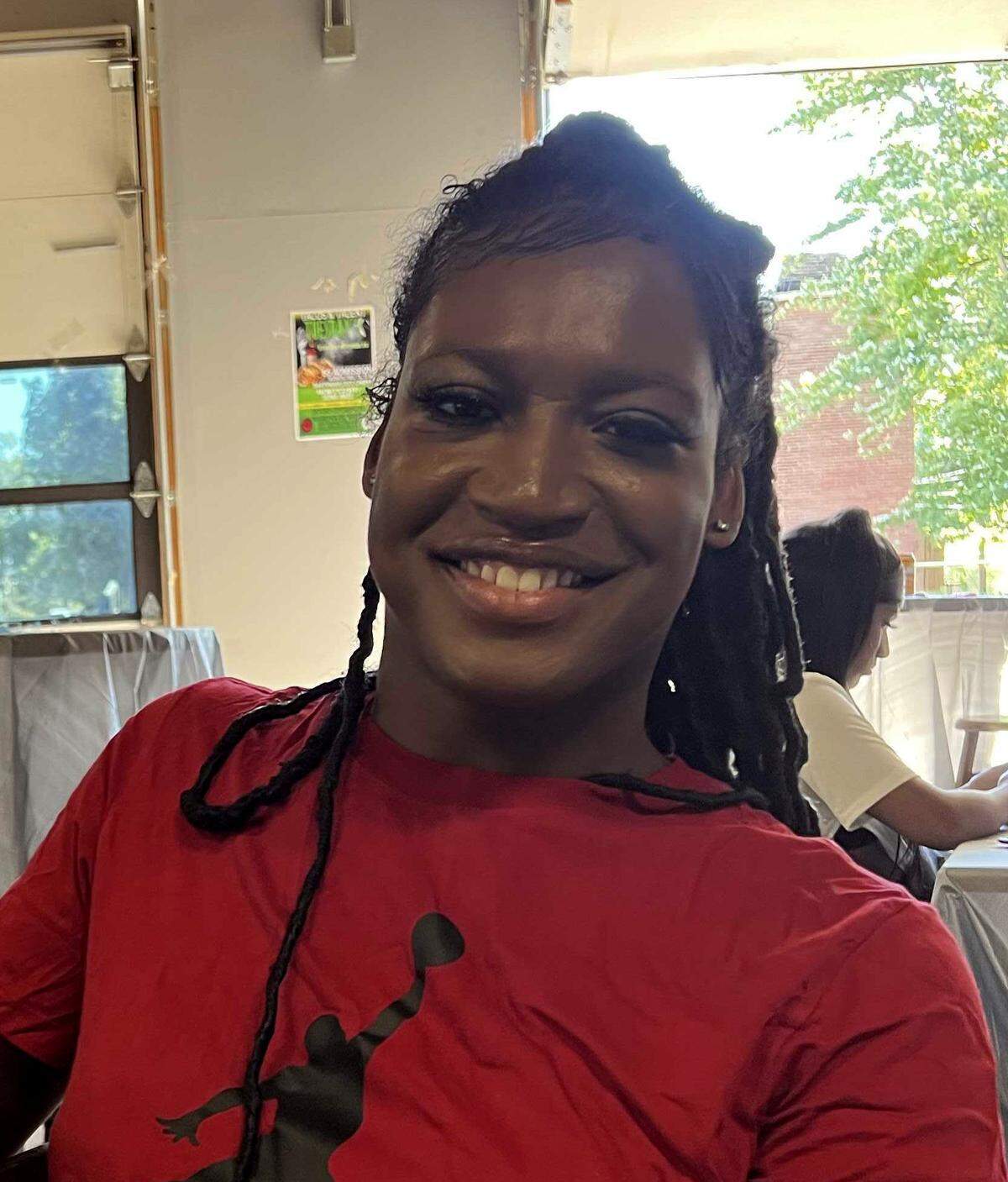 A Black transgender woman, wearing a red shirt, smiles for a photo.