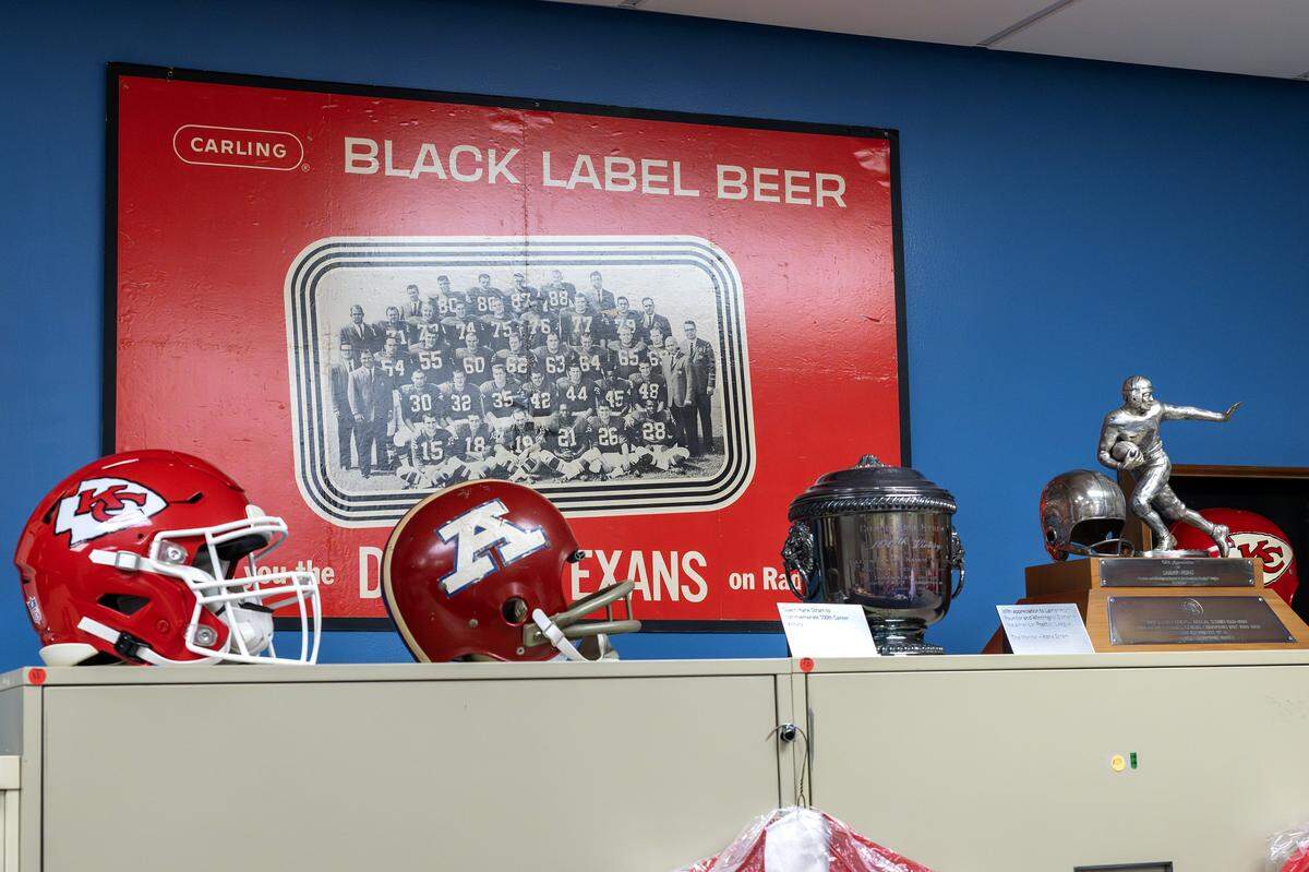 Kansas City Chiefs and Dallas Texans helmets are displayed alongside team trophies and memorabilia inside the Library Room at the team’s underground storage facility  in Kansas City. A vintage Carling Black Label Beer poster featuring the 1962 Dallas Texans team hangs in the background.