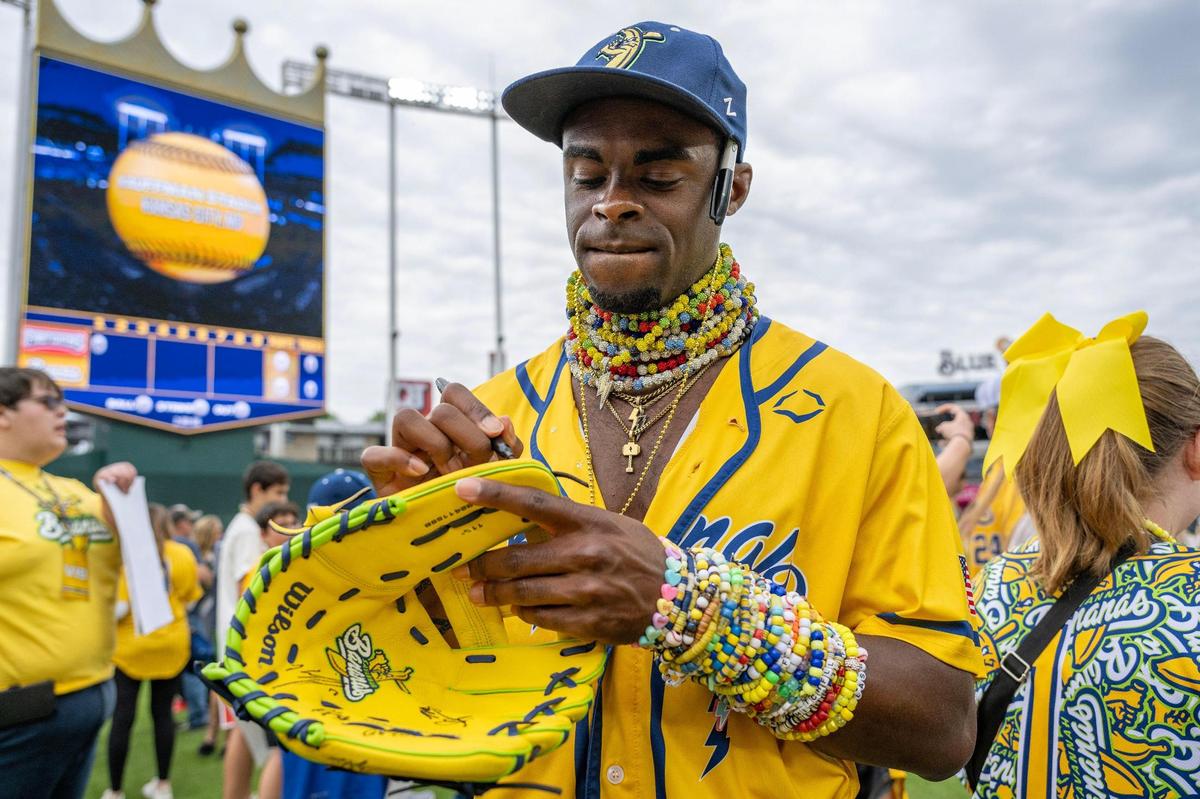 Savannah Bananas designated runner Malachi Mitchell, wearing multiple necklaces and bracelets, signs a baseball glove during VIB fan access before a Banana Ball game against the Firefighters at Kauffman Stadium on Friday, May 23, 2025, in Kansas City.