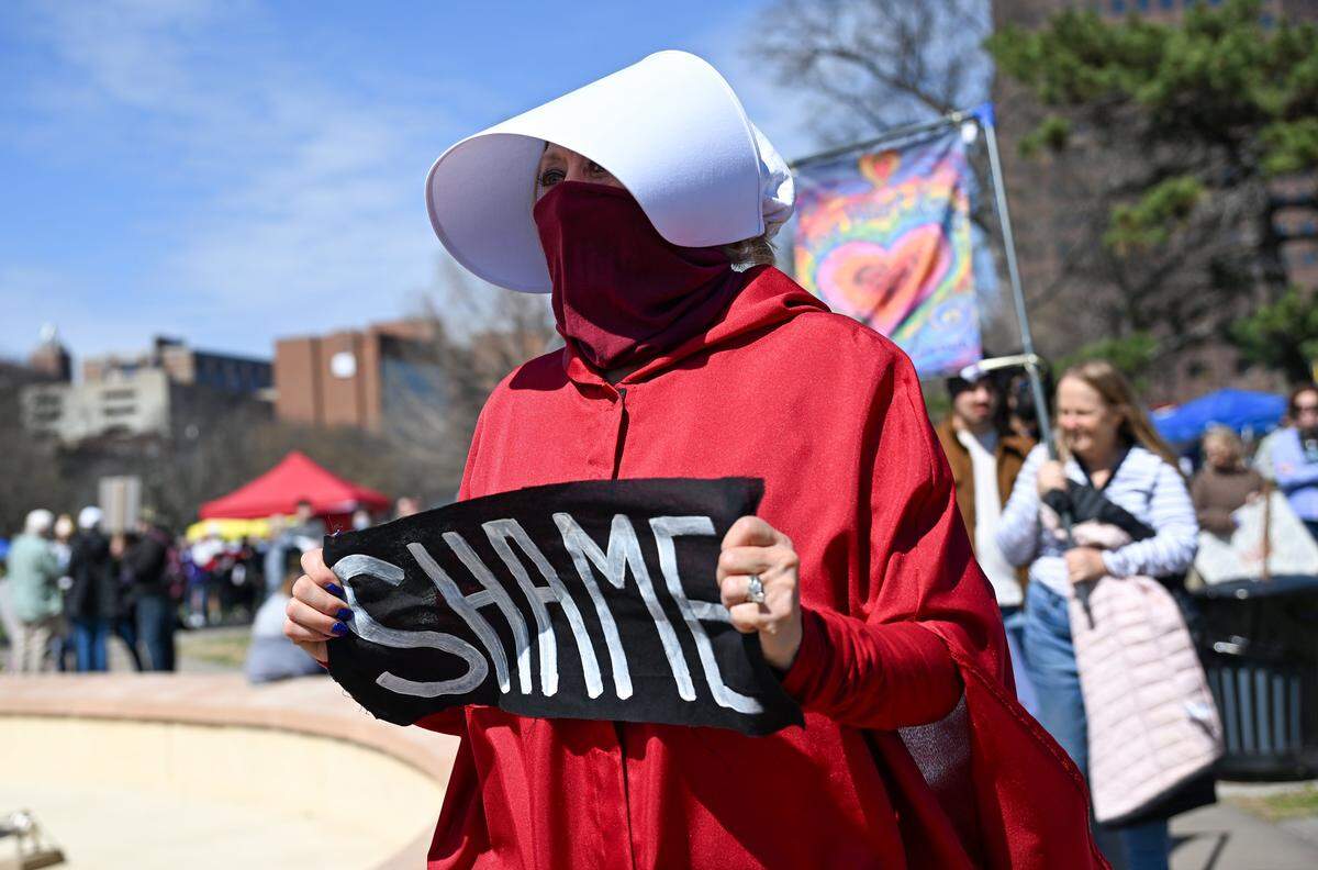 A woman dressed in garb from “The Handmaid's Tale” holds up a sign in Mill Creek Park near the Country Club Plaza on Saturday, March 28, 2026, in Kansas City for a No Kings protest against the Trump administration.