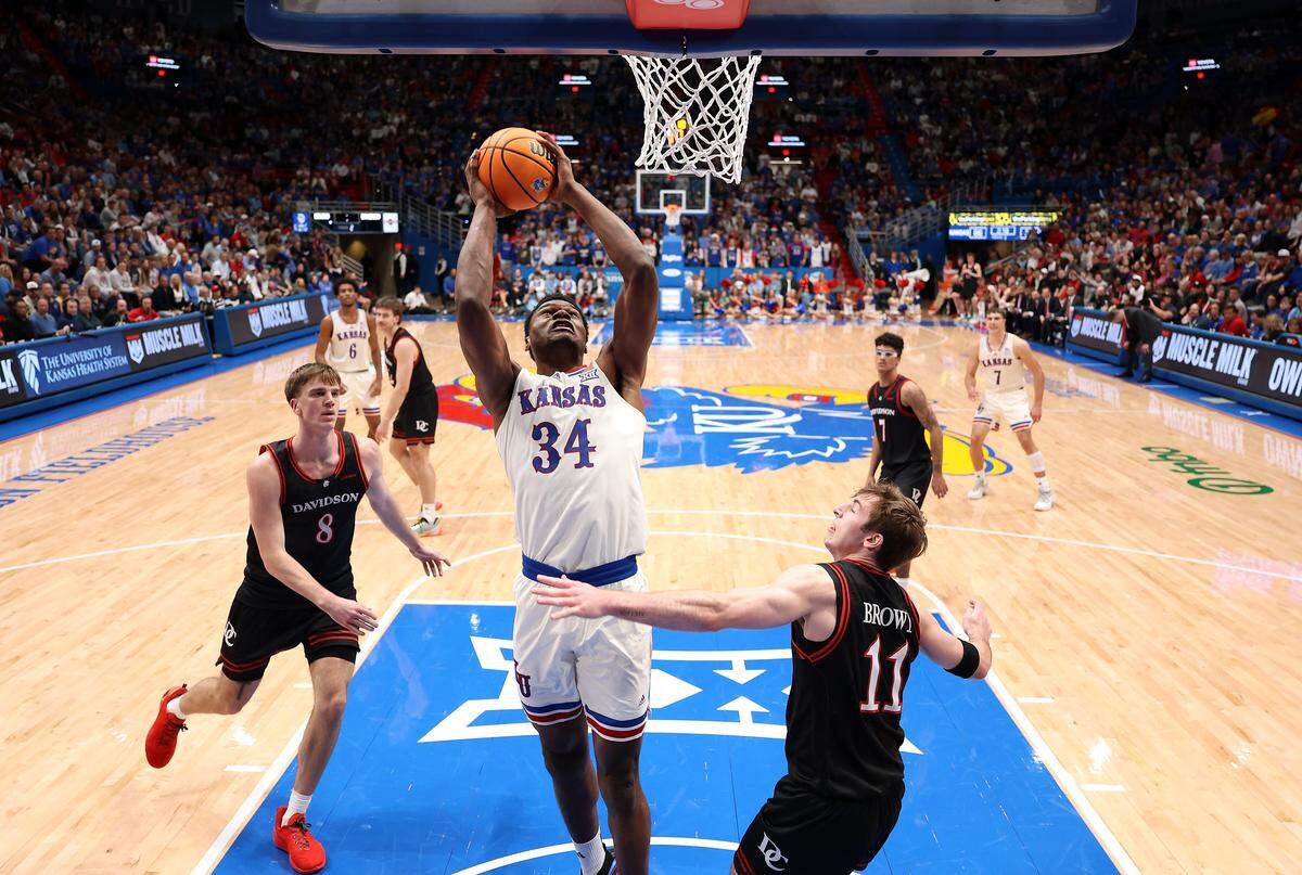 Paul Mbiya #34 of the Kansas Jayhawks attacks the basket as Joe Hurlburt #8 and Sam Brown #11 of the Davidson Wildcats defend during the game at Allen Fieldhouse on December 22, 2025 in Lawrence.