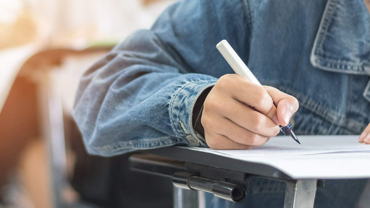 Photo illustration of a student’s hand holding a pen while writing on a piece of paper at a school desk