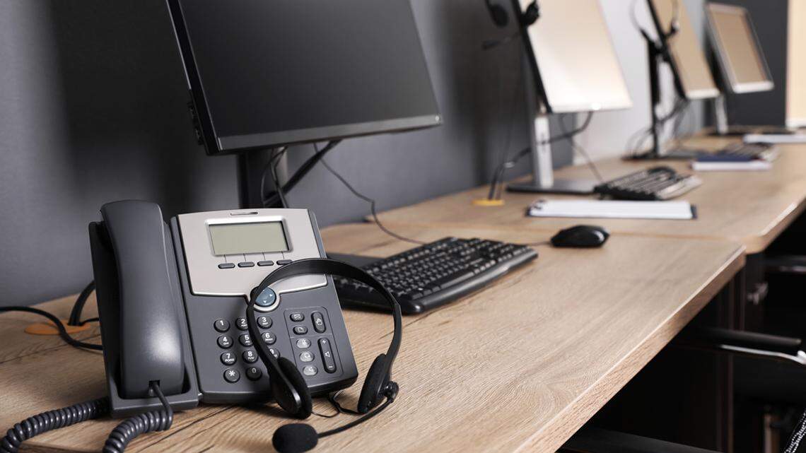 Stationary phone and headset near modern computer on wooden desk in office. 