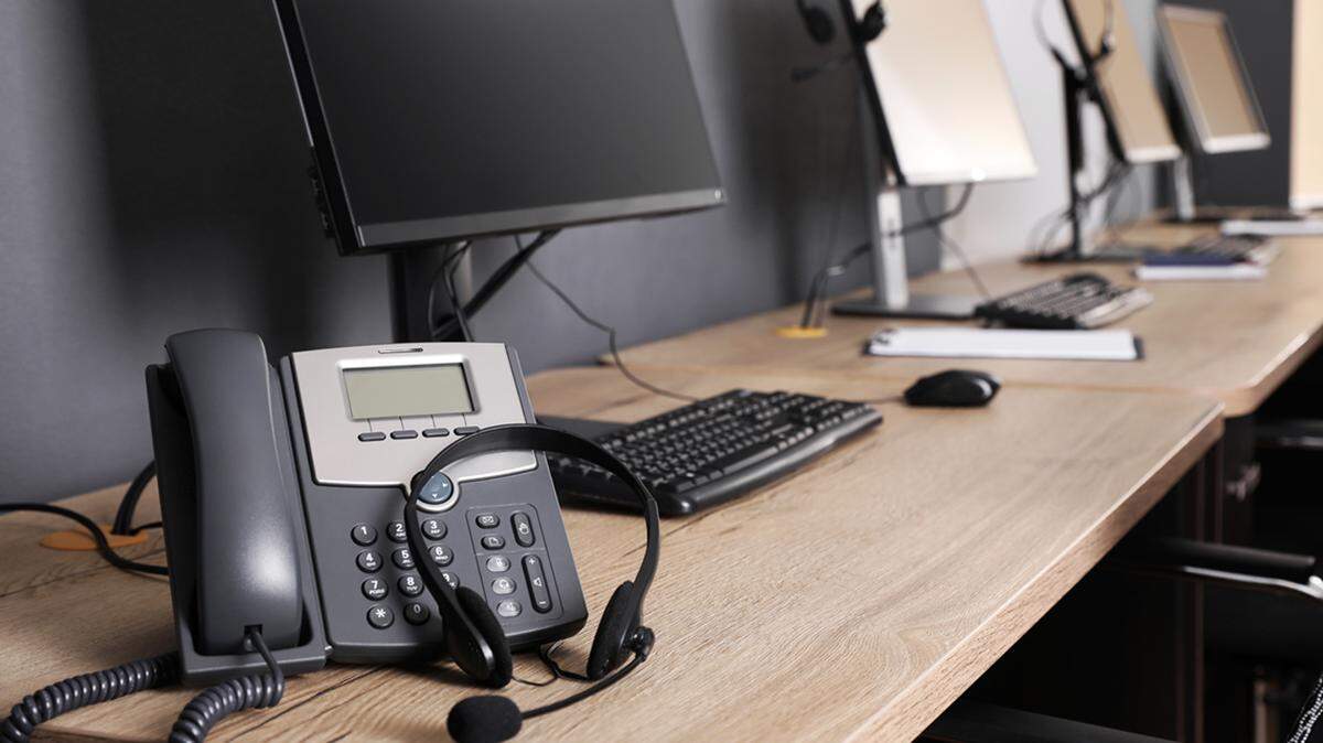 Stationary phone and headset near modern computer on wooden desk in office. Hotline service telephone call center suicide prevention help line