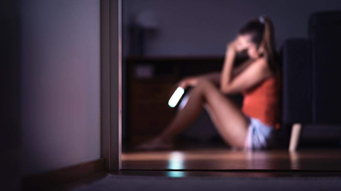 Sad teenager with smartphone in her room sitting on floor. Lonely teen girl crying with mobile phone. Harassment or online cyber bullying in social media or internet. Mean message or troll comment.