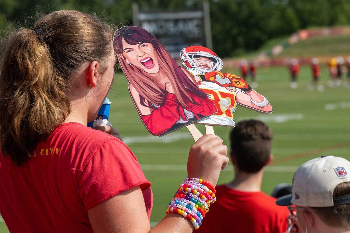 A young Chiefs fan wearing a wristful of friendship bracelets holds cardboard cutouts of Taylor Swift and Travis Kelce during practice at Chiefs training camp.