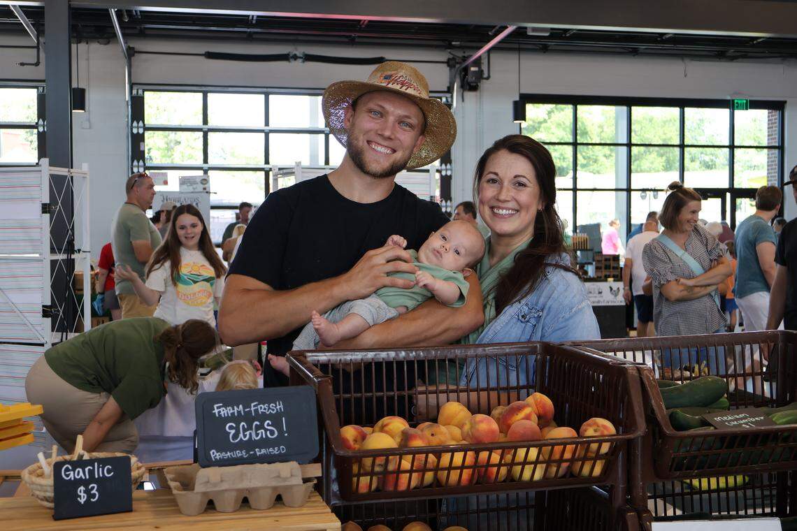 Two vendors and their baby smile behind their stand at the farmers market.