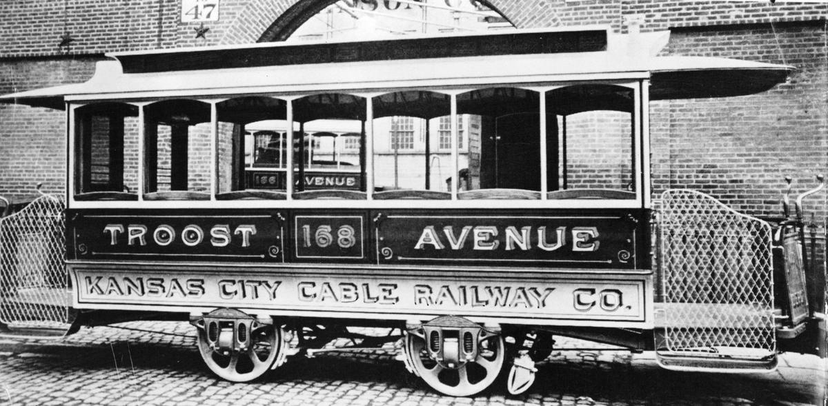 Typical of the cable car trailers put into use in Kansas City in the 1880s was this one on the Troost line. It was towed by a grip car which was propelled by a moving underground cable. The gates were removed to expedite loading and unloading. Except at major stops, men usually boarded or hopped off without waiting for the cars to come to a halt.