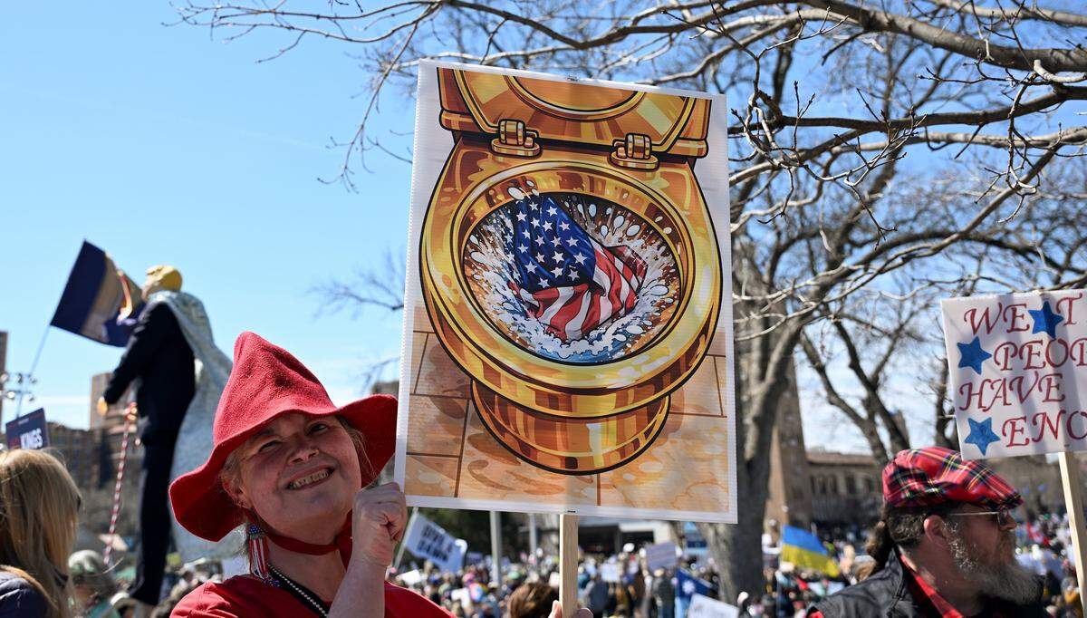 A protester holds a sign depicting a golden toilet flushing an American flag during a No Kings protest at Mill Creek Park near the Country Club Plaza on Saturday, March 28, 2026, in Kansas City.