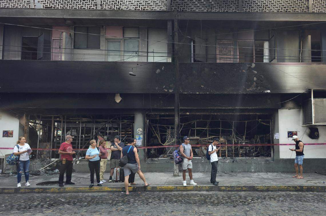People stand next to a burned building in Puerto Vallarta, Jalisco state, Mexico, on February 23, 2026. Mexico has deployed 10,000 troops to quell clashes sparked by the killing of the country's most wanted drug lord, Nemesio "El Mencho" Oseguera, leader of the Jalisco New Generation Cartel (CJNG).