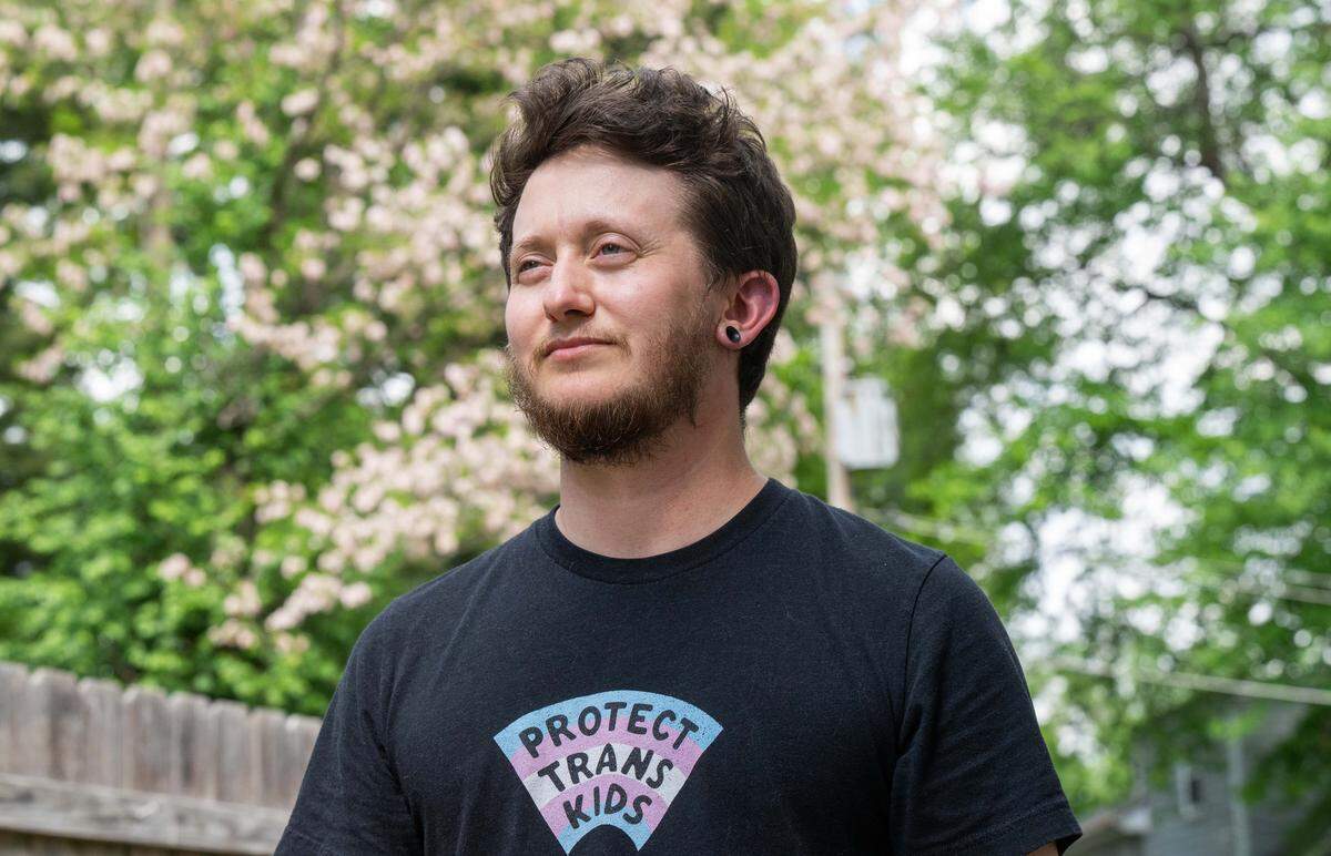 Riley Long stands with trees in the background. He wears a shirt with the words “protect trans kids.”