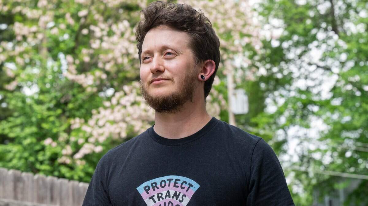 Riley Long stands with trees in the background. He wears a shirt with the words “protect trans kids.”