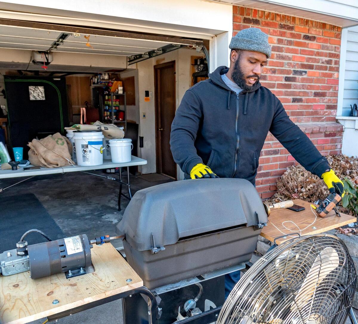 Brent Rice and a friend modified an ordinary propane grill to create a coffee bean roasting setup that he fires up in his driveway. The table behind him holds raw, organic coffee beans and containers of already roasted beans that are cooling.