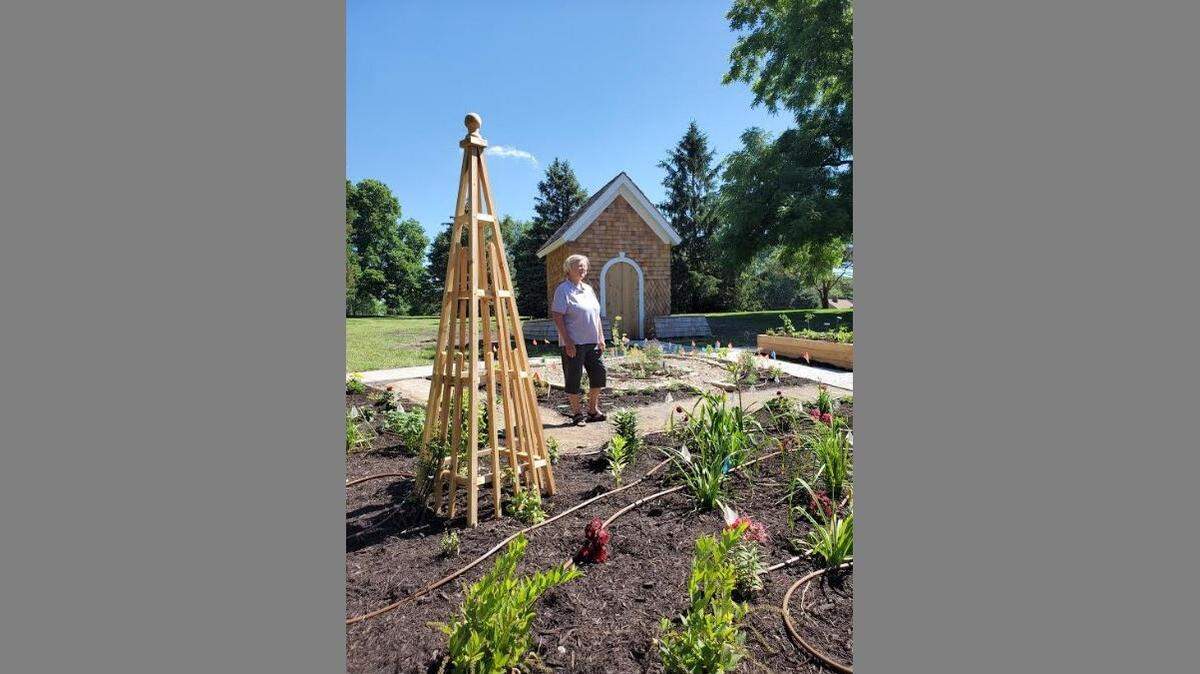 Shawnee resident Cindy Hobbs, one of three Extension Master Gardeners spearheading the West Flanders Park project, stands in front of a wooden cottage-like structure — built by local contractor Henry Specht — to house gardening tools and equipment.
