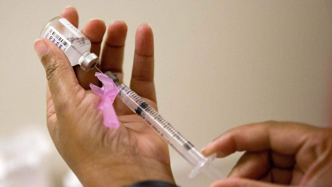 A nurse's left hand holds a vial filled with the seasonal influenza vaccine while her right hand uses a needle and syringe to draw a single dose into the syringe.