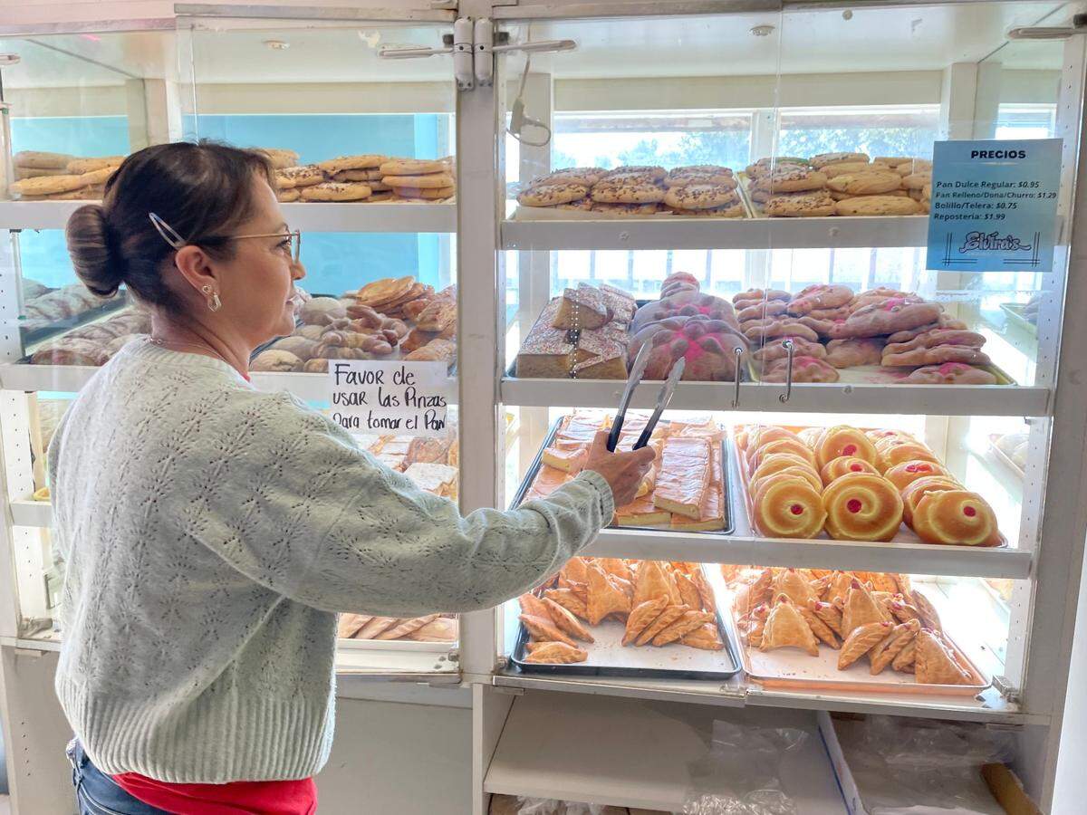 Elvira Arizmendi, owner of Elvira’s Cakes stand in front of bread cabinet with tongs pointing to traditional pan de muerto for the Day of the Dead.