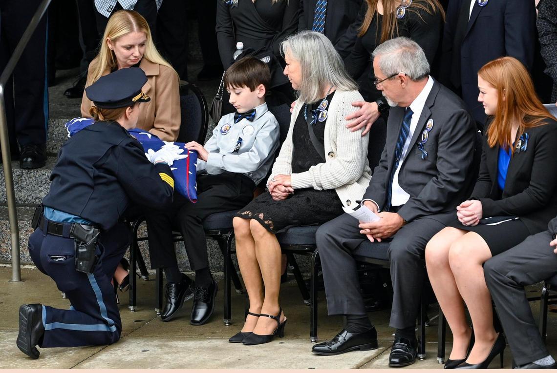 Kansas City Police Chief Stacey Graves, left, presents a flag to Ayden, a son of fallen Kansas City Police Officer James Muhlbauer, 42, during the funeral for Muhlbauer and his police K-9, Champ, Wednesday, Feb. 22, 2023, at Municipal Auditorium in Kansas City. Muhlbauer’s family, including his wife, Cassie, left, looks on.