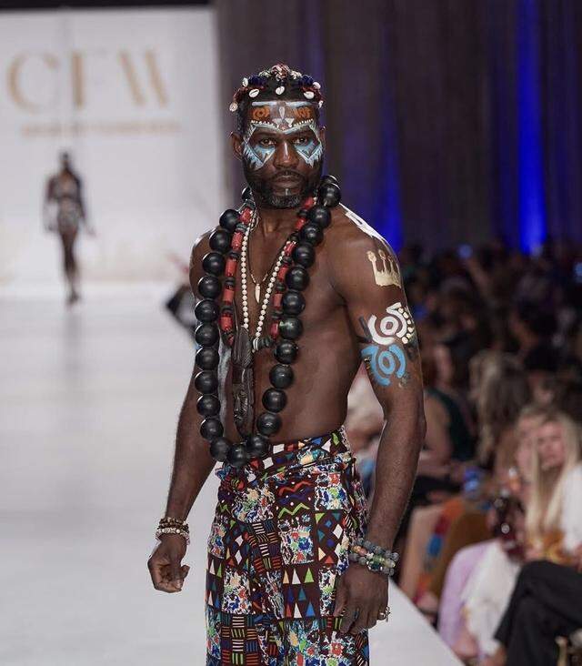 A male model Adeleye Arasanyin wears outfit designed by Kenya Martin with pants made from African-inspired patchwork designs with large wooden bead necklaces atop his bare chest along with body paint adorning his arms and face.