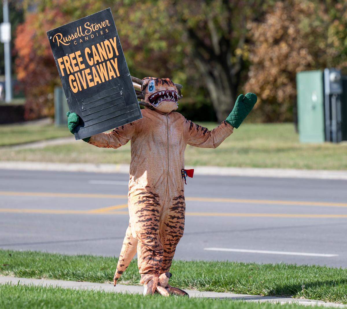 A Russell Stover employee dressed in a dinosaur costume waves a sign promoting the company's free candy giveaway outside the Russell Stover headquarters on Friday, Oct. 31, 2025, in Kansas City.