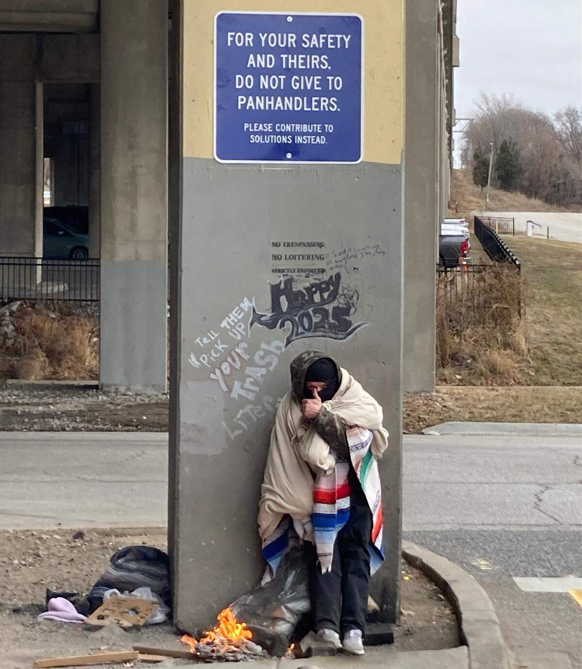 Thomas Gonzales, 38, huddles in the cold beneath a blue sign, recently affixed at the underpass of Interstate 35 where the highway exits onto West Pennway Street. The sign urges people not to give money to panhandlers. 