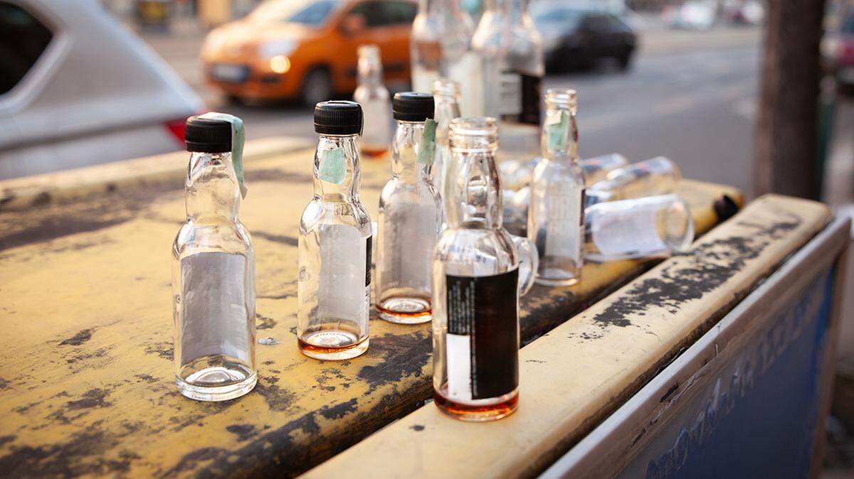 Selective-focus street view of empty miniature liquor bottles scattered across the scratched lid of a weathered kiosk in central Budapest. Clear glass vials with black caps hold a few drops of whisky and rum, while others lie on their sides awaiting collection by sanitation crews. Background cars and buildings dissolve into creamy bokeh, contrasting morning calm with the gritty evidence of last night’s binge drinking. The photograph raises issues of alcohol culture, public-space littering, recycling habits and the social cost of nightlife in European capitals where open-container rules are lightly enforced.