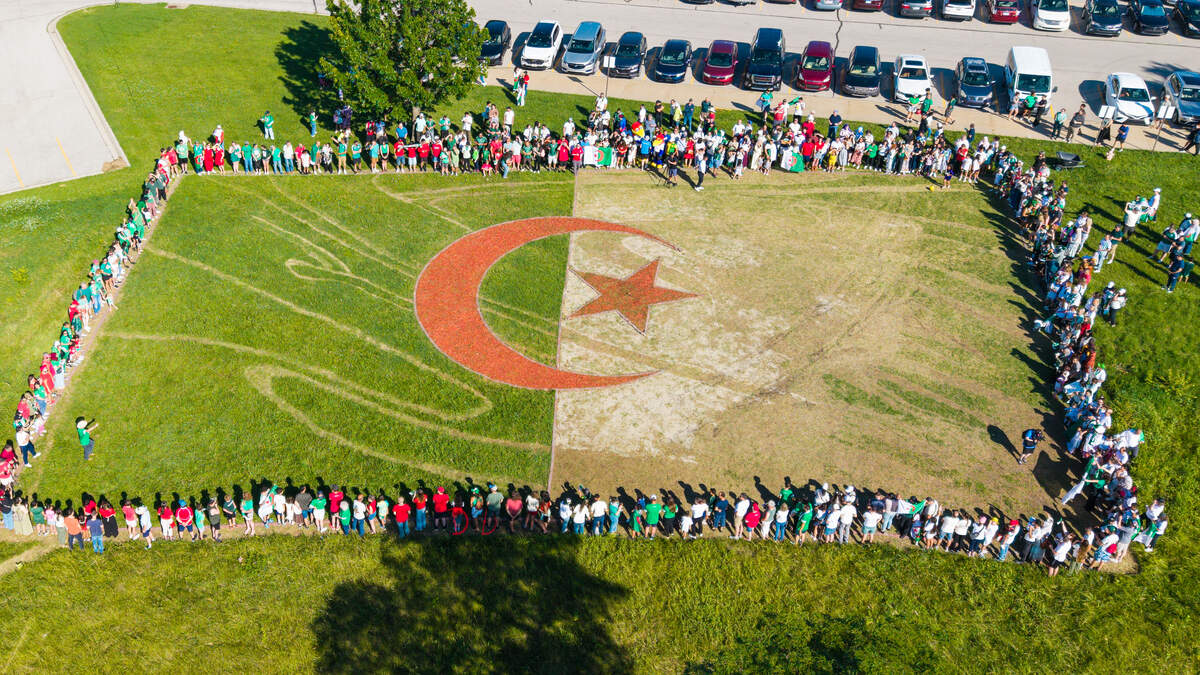 A crowd starts to gather around earthwork artist Stan Herd’s grassy creation of a billowing Algeria flag on June 13, 2026. The crowd would grow to over 800 into the evening during a block party event held onsite — food, music and celebration of a town accepting Algeria wholeheartedly brought Herd to tears.